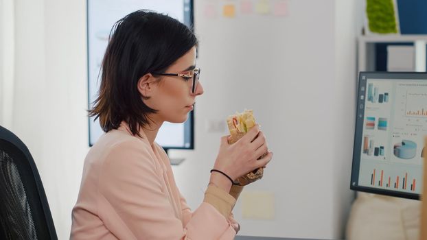Entrepreneur woman eating tasty sandwich having meal break working in business company office during takeout lunchbreak. Fast food order paper bag delivered at workplace. Manager typing strategy