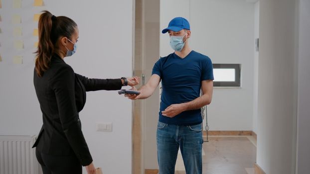 Delivery guy with medical face mask and gloves against coronavirus delivering takeaway food meal order in company office. Businesswoman paying lunch with smart watch using POS contactless service