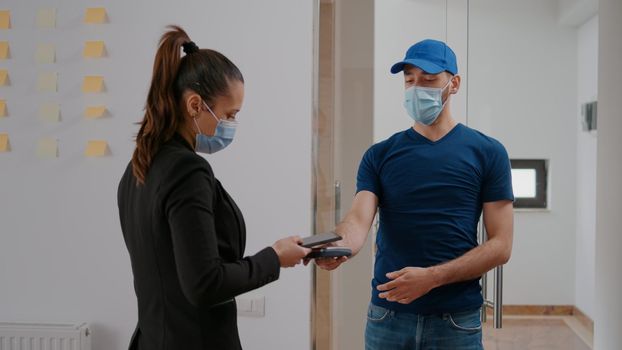 Delivery guy with protection face mask holding POS terminal delivering takeaway food order bag to entrepreneur client during lunchtime. Businesswoman paying meal service using contactless smartphone