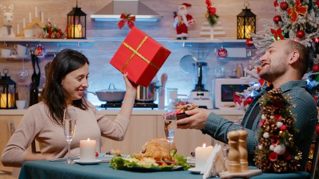 Man and woman celebrating christmas at festive dinner, enjoying holiday festivity while exchanging presents decorated with bow, ribbon and wrapping paper. Couple giving gifts for celebration