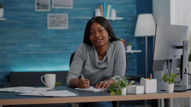 Point of view of student with dark skin smiling at camera during communication videocall telework conference meeting. African american woman sitting at desk in living room working remote from home