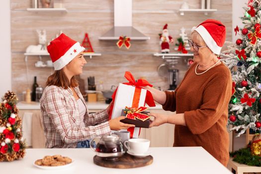 Grandmother surprising granddaughter with xmas wrapper gift celebrating christmas holiday together in decorated kitchen. Child enjoying winter season changing present with ribbon on it with gradma