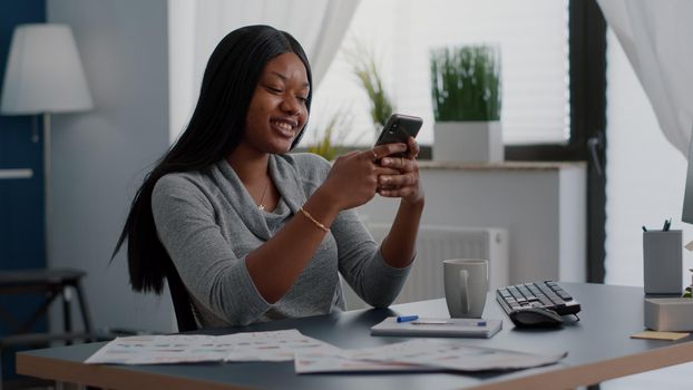 Student with black skin texting message using modern phone while sitting at desk working remote from home. African american woman looking on social media sharing lifestyle advice chatting with friends