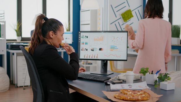 Closeup of businesswoman eating tasty pizza while analyzing financial statistics at desk in workplace. Takeout order food delivery in corporate job place, lunch meal break package delivered at startup office.