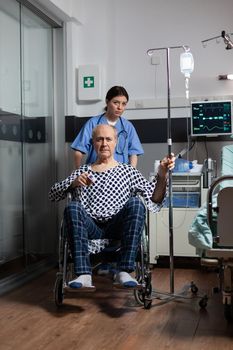 Medical practitioner preparing senior sick hospitalized man in wheelchair, checking iv drip. Nurse with stethoscope preparing patient for examinantion. Oximeter attached to finger of man.