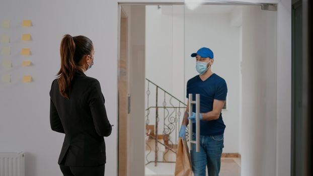 Businesswoman with face mask against coronavirus paying takeaway food order with smartphone using POS contactless service. Delivery guy with gloves delivering restaurant meal bag during lunchtime