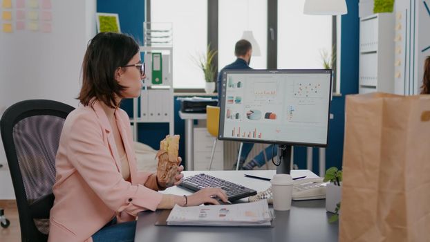 Businesswoman eating tasty sandwich having meal break working in business company office during takeout lunchbreak. Fast food order paper bag delivered at workplace. Woman typing strategy on computer