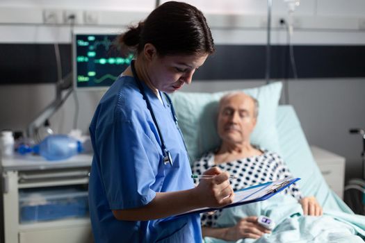 Medical assistant checking treatment of senior man reading notes on clipboard. Sick elderly laying in bed discussing with nurse about diagnosis expertise with iv bag attached.