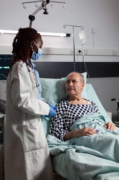 African american medical practitoner wearing chirurgical mask, in hospital room during consultation of sick unwell senior man, breathing with help from test tube and iv drip attached.