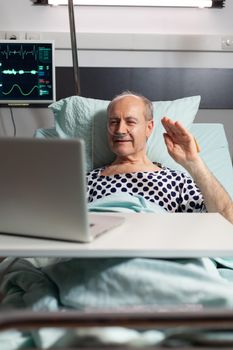 Cheerful sick senior man waving at camera during video conference using laptop laying in bed, breathing through oxygen tube. Man recovering after surgery and treatment.