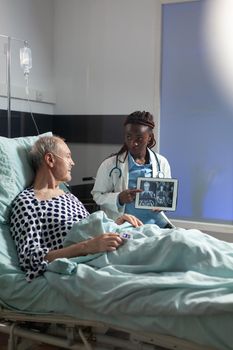 African american therapist showing x-ray on tablet pc discussing explaining diagnosis and treatment to sick old man breathing with oxygen tube laying in bed.