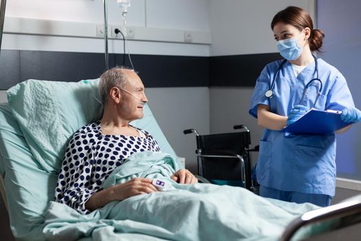 Medical nurse in scrubs with chirurgical mask taking notest on cliboard during consultation of sick unwell senior man. Patient with iv drip attached breathing with help from oxygen mask.