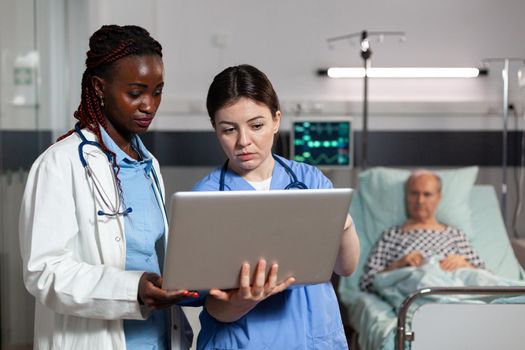 African american doctor and nurse using laptop in hospital room for sick senior patient diagnosis and treatment. Old man laying in bed with iv drip attached breathing through oxygen mask.