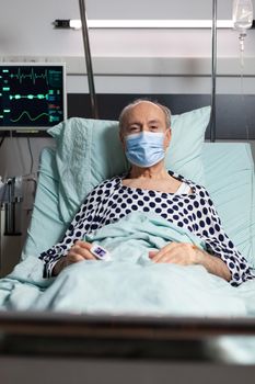 Portrait of sick senior man patient with chirurgical mask resting in hospital bed, with iv drip attached on hand having oximeter attached on finger. Modern medical equipment.