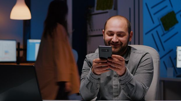 Entrepreneur man smiling at phone while writing financial strategy planner sitting at desk table in company office. Businessman browsing social media ideas on internet late at night