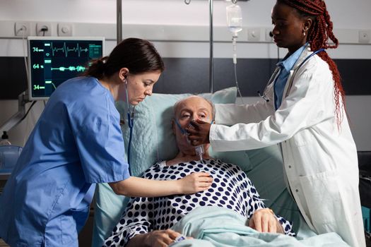 African doctor and medical assistant helping senior man breath using oxygen mask, in hospital laying in bed. Sick patient cant breath because of lung infection. Nurse listening heart with stethoscope.