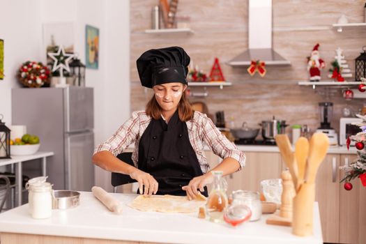 Child wearing baking apron putting flour on homemade dough preparing traditional gingerbread dessert enjoying winter season in xmas decorated kitchen. Kid celebrating christmas holiday making cookies