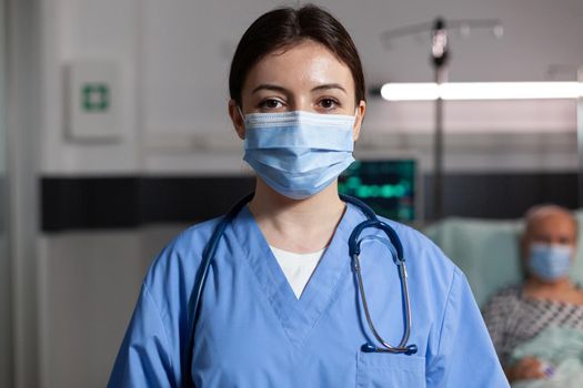 Doctor health care specialist wearing chirurgial mask in hospital room looking at camera with sick patient breathing with help from oxygen tube and iv drip attached.