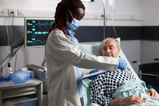 African american medical practitoner wearing chirurgical mask, in hospital room during consultation of sick unwell senior man, breathing with help from test tube and iv drip attached.