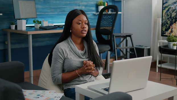 Student with dark skin waving school team working remote from home at marketing course during online videocall telework conference meeting. Computer user sitting on bean bag in living room