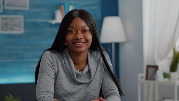 Portrait of confident student relaxing at desk table in living room looking into camera smiling during homeschooling. Black teenager browsing high school information working at academic books