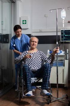 Hospitalized senior man sitting in wheelchair in hospital room, holding iv drip with oximeter attached on finger. Medcal assistant preparing sick patient for treatment.