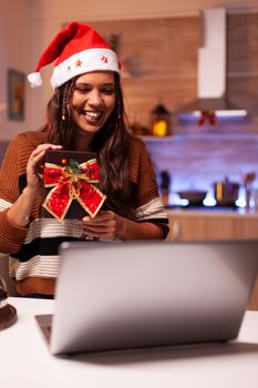 Joyful woman holding present box while on video call conference concept with friends in festive kitchen. Caucasian young adult receiving surprise gift for christmas eve seasonal festivity