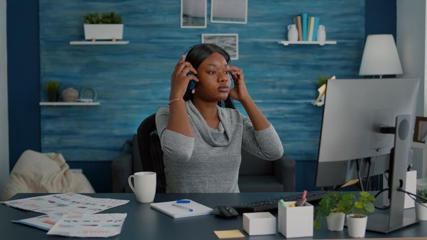 Student with black skin putting headphones listening music browsing school ideas on internet using e learning platform on computer. Young woman sitting at desk table in living room enjoying fun break