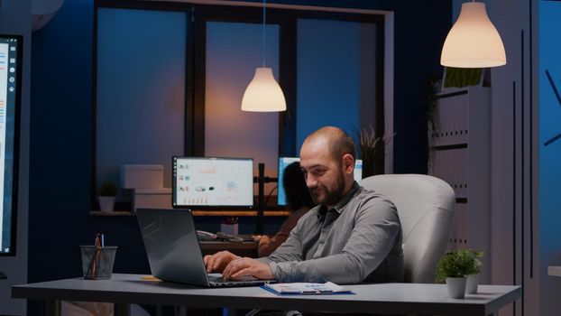 Exhausted businessman checking marketing statistics on laptop computer working in startup company office. Tired workaholic manager remains alone in financial room after his collegue left