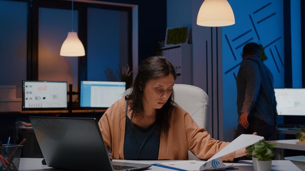 Overworked businesswoman analyzing financial graphs on clipboard comparing with economic statistics using laptop computer. Tired entrepreneur woman working in business company office late at night