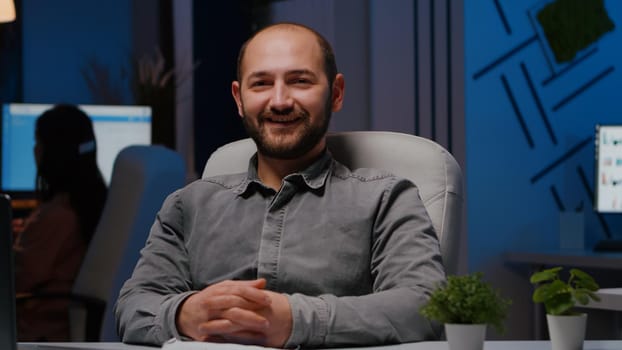 Portrait of smiling businessman sitting at desk table in business company office late at night looking into camera. Executive manager working at marketing statistics planning financial strategy