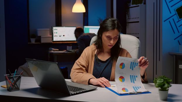 Exhausted workaholic businesswoman checking marketing strategy on clipboard in startup office late at night. Overworked executive manager working on financial graphs company deadline