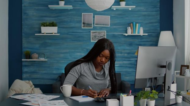 Student writing business ideas on sticky notes putting on computer working at school homework, using elearning platform during university online course. Black woman sitting at desk in living room