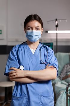 Portrait of medical nurse with chirurgical mask in hospital room with sick unweel senior pantient with trauma looking at camea with arms crossed, wearing scrubs.