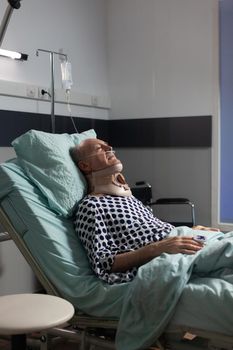 Senior man suffering after serious accident laying in hospital bed, wearing neck brace. Patient with cervical collar unconscious with iv drip attached and oximeter on finger waiting for treatment.