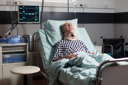 Elderly man laying in hospital room bed wearing cerival collar, with iv drip. Oxygen mask helping patient breath in intensive care clinic. Sleeping hospitalized man with neck trauma.