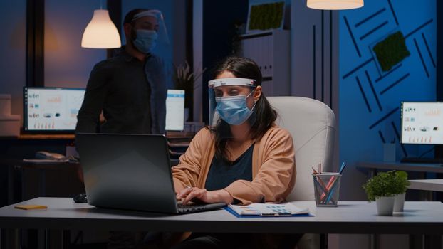 Workaholic tired businesswoman with face mask and visor against covid19 working in company office typing financial statistics on laptop. Manager remains alone in company room after collegue left