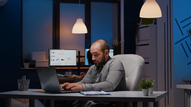 Overworked businessman typing management strategy on laptop working in startup company office late at night. Tired exhausted manager he left alone in corporate room analysing economic statistics