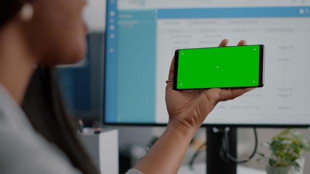 Black woman looking at isolated display having online videocall conference meeting using mock up green screen chroma key phone sitting at desk in living room. Student watching social media video