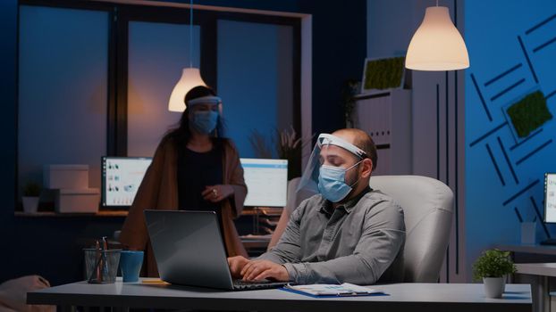 Workaholic businessman with face mask and visor against covid19 working in company office analysing marketing statistics on laptop. Manager remains alone in company room after his collegue left