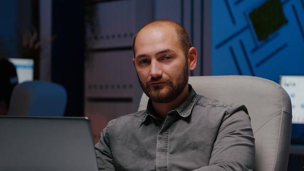 Portrait of exhausted workaholic man manager typing financial strategy using laptop computer while sitting at desk table in business company office. Tired businessman looking into camera late at night