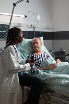 African american therapist in hospital room discussing treatment holding pills bottle reassuring recovery,sick senior man laying in bed breathing through oxygen mask,