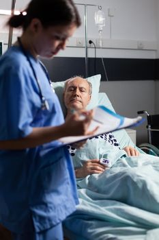 Medical nurse in scrubs taking notest on cliboard after consultation of sick unwell senior man. Patient with iv drip attached breathing with help from oxygen mask.