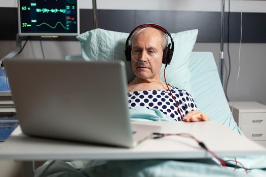 Senior sick man in hospital bed breathing through oxygen mask, listening music on headphones, looking at laptop. Old man monitored by modern equipment.
