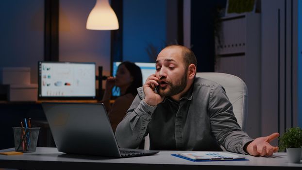 Stressed angry businessman sitting at desk table discussing corporate problem at phone having breakdown late at night in startup company office. Unhappy nervos frustrated manager resolving job problem