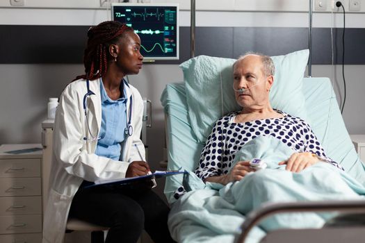 African american doctor reading diagnosis from clipboard to sick ill unwell senior patient laying in bed, breathing with help from oxygen mask, listening discussing with medical staff about recovery.