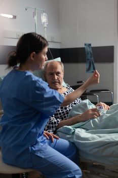 Medical nurse analyzing senior patient chest x-ray in hospital room, discussing explaining diagnosis. Old man breathing through respiratory tube laying in bed listening health care specialist.