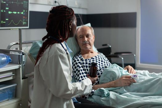 Senior man breathing with help from oxygen tube laying in hospital bed, listening african american doctor explaying diagnosis treatmant holding pills bottle for recovery.