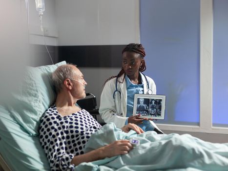 African american doctor sitting next to senior man explaining body trauma diagnosis, showing x-ray on tablet pc in hospital room. Mature patient in clinic breathing it help from oxygen mask.