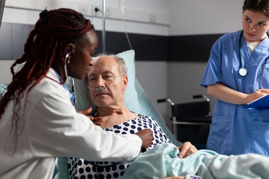 African american cardiologist checking examining senior patient heart, using stethoscope while patient is laying in hospital bed to set diagnosis for therapy, Breathing with help from test tube.